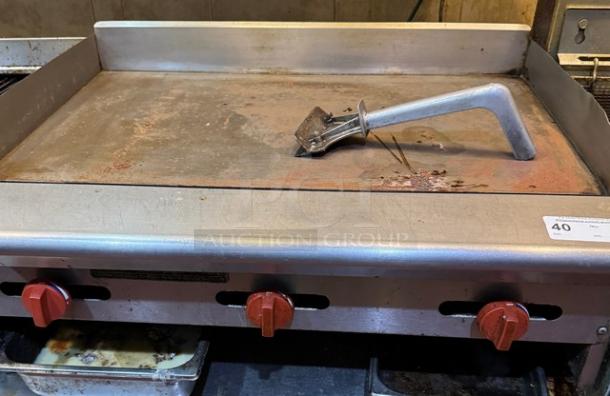 36" flat top gas griddle with red controls and metal scraper; tested, working, some surface wear; ideal for commercial use.