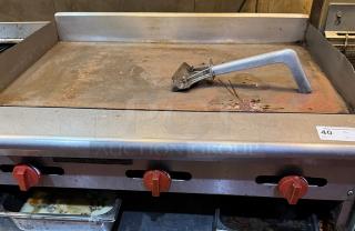 36" flat top gas griddle with red controls and metal scraper; tested, working, some surface wear; ideal for commercial use.