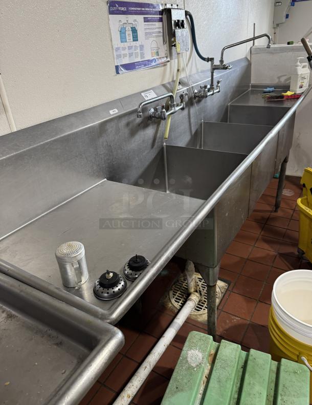 Stainless steel 3 compartment sink with 2 drain boards, multiple faucets, and wall-mounted instructions, in used condition.