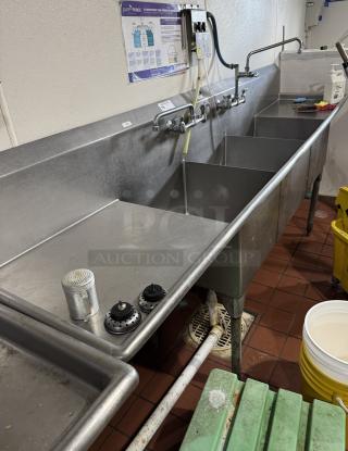 Stainless steel 3 compartment sink with 2 drain boards, multiple faucets, and wall-mounted instructions, in used condition.