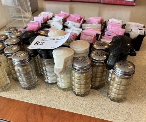 Assorted salt and pepper shakers with metal lids and sugar packet holder, in good condition, labeled "82" for auction.
