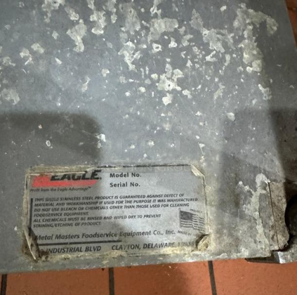Eagle work table with weathered stainless steel surface, under shelf, and manufacturer label showing brand and serial details.