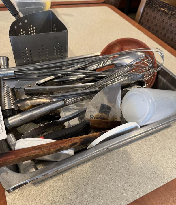 Assorted kitchen utensils and pasta strainer in a metal container. Includes tongs, whisks, and a grater. Mixed materials, used condition.