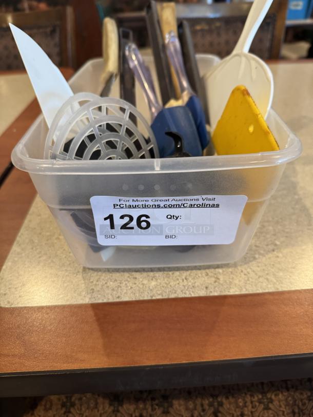 Assorted kitchen utensils in a clear plastic bin, item number 126. Includes spatulas, spoons, and a strainer.
