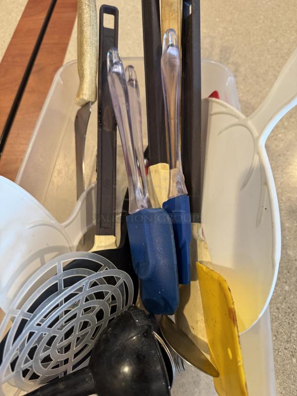 Assorted kitchen utensils in a plastic container, including spatulas, ladles, and tongs. Various colors and materials, gently used.