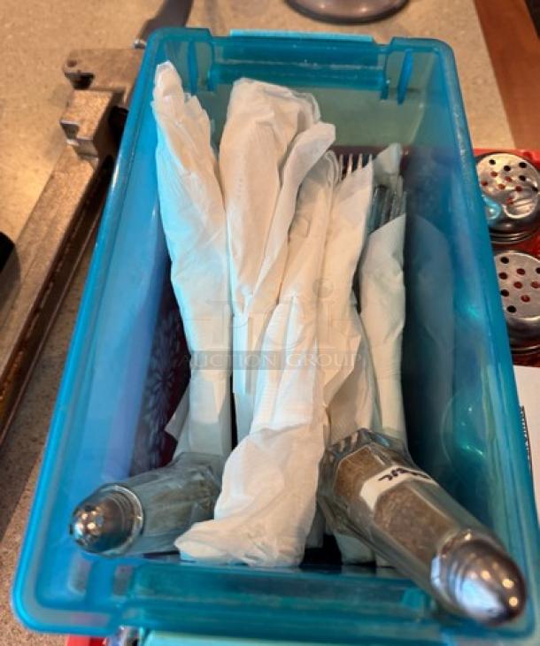 Assorted shakers and wrapped flatware in a blue container; shakers contain spices with visible labels, flatware wrapped in napkins.