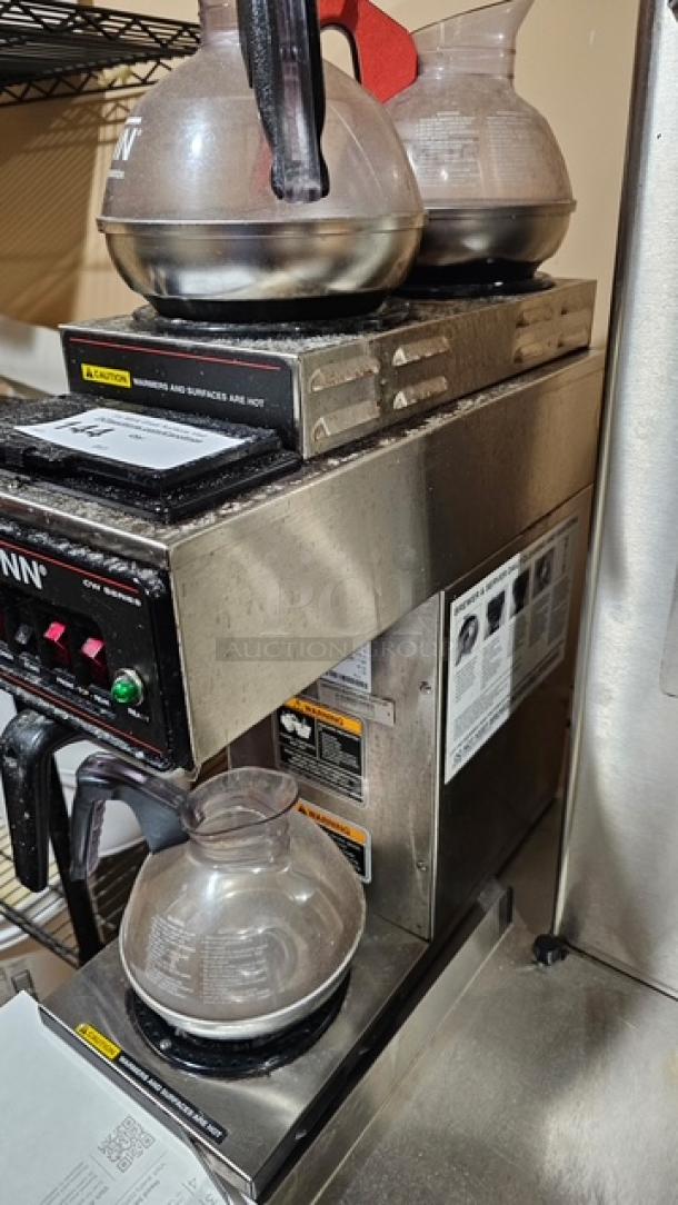 Bunn coffee maker with dual top warmers and three glass pots. Stainless steel exterior with visible control panel and safety labels.