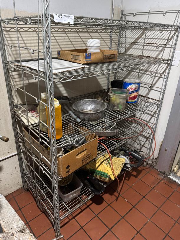 Metal wire floor rack with five shelves, used condition, containing various items. Label marked "105" on the top shelf.