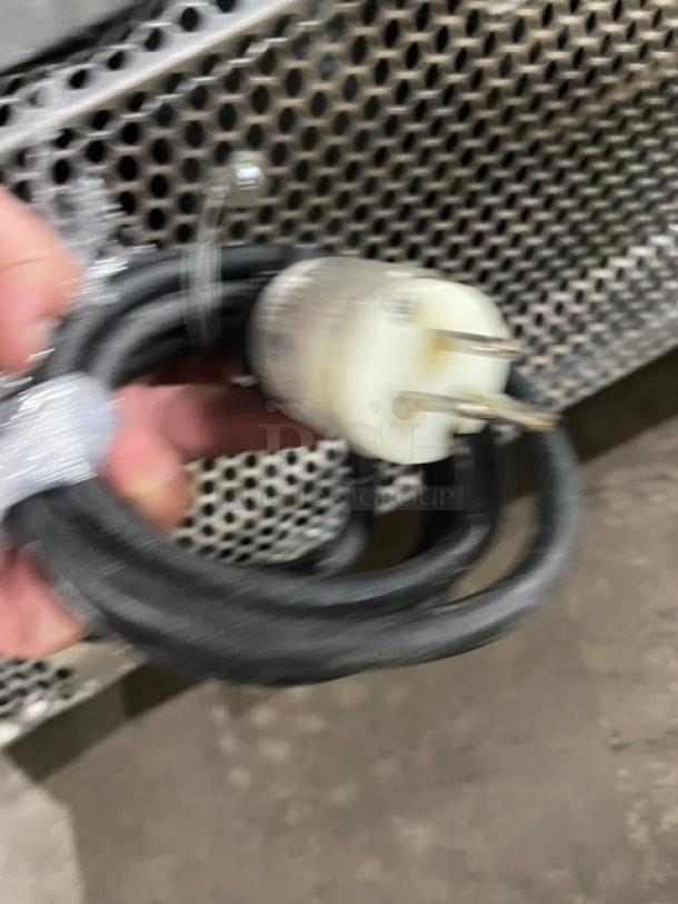 Blurry image of a power cord and plug held in front of a perforated metal surface, likely part of an appliance or machinery.