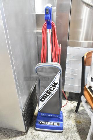 Brand new Oreck XL2100RHS upright vacuum, 12" wide, lightweight, bagged, with visible scratches and dents. 120V, 1 phase.