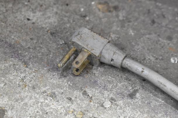 Close-up of a worn electrical plug on a textured surface. The plug appears to be for a commercial appliance.