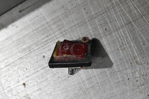 A small broken red switch on a scratched metal surface, possibly detached from a larger appliance.