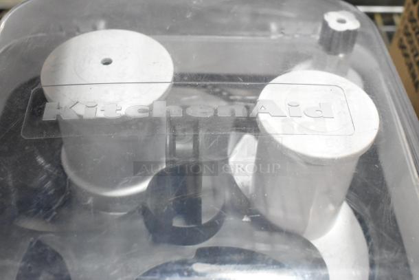 KitchenAid food processor parts under clear plastic lid, featuring cylindrical components. Brand marked on lid. Scratch and dent condition.