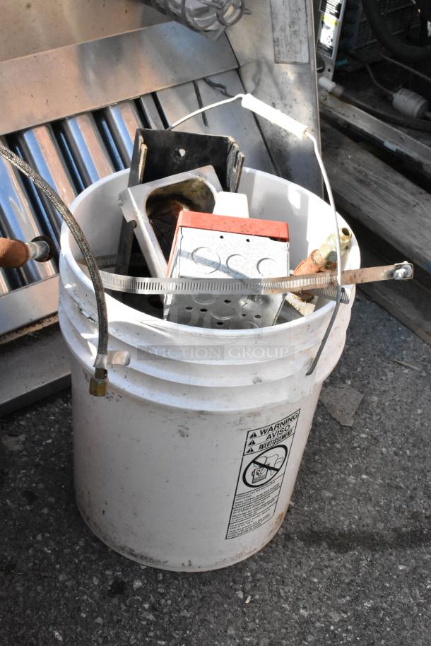 White bucket containing metal parts and hoses; includes warning label. Background shows metal commercial kitchen equipment.