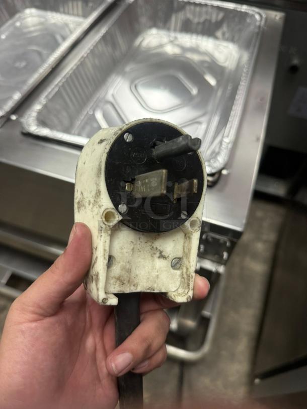 A worn electrical plug with visible dirt, held in hand. Features three prongs, black and white casing, screws in place.