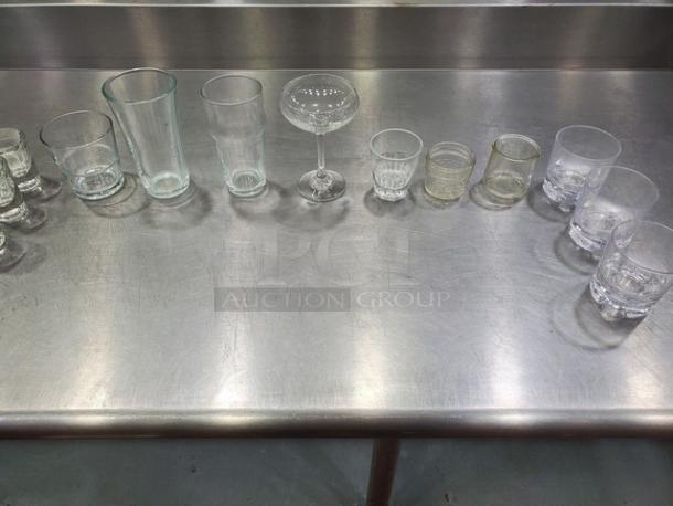 Lot of 26 miscellaneous glasses, including tumblers, goblets, and shot glasses; varied designs on stainless steel surface.