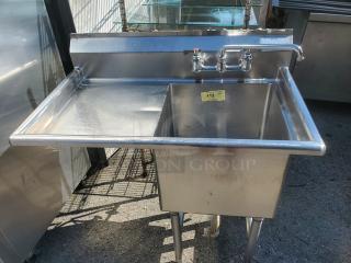 Stainless steel one compartment sink with integrated backsplash, side drainboard, and dual-handle faucet. Auction tag "112".