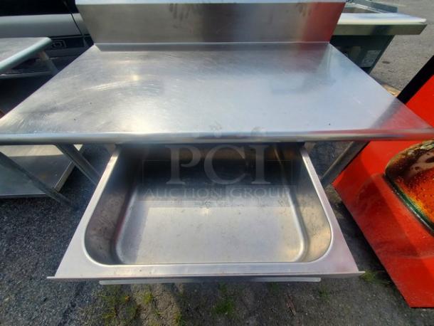 Stainless steel work table with drawer, medium size, set outdoors. Shows signs of use on metal surface.