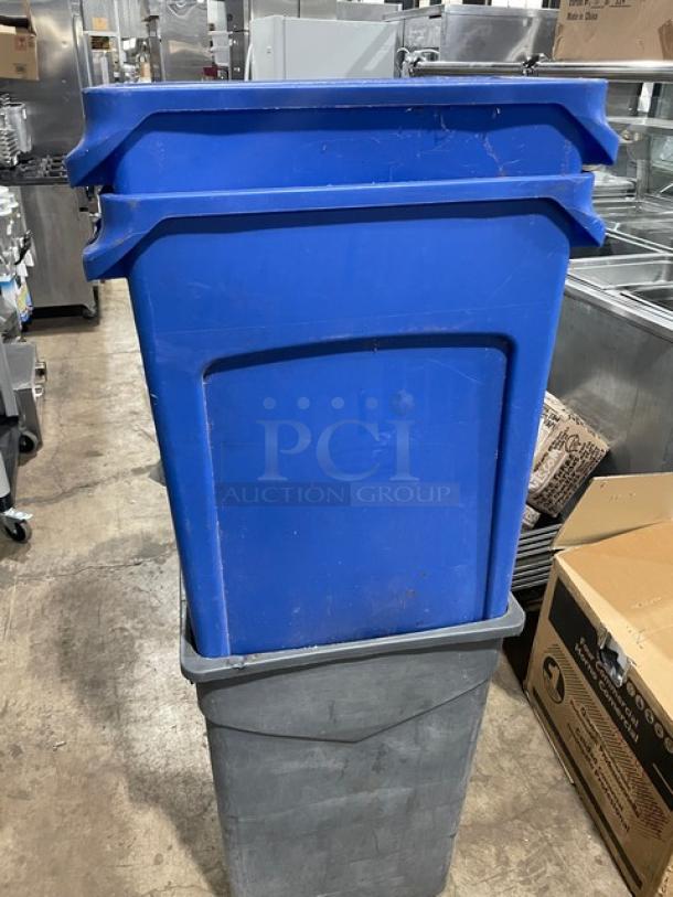 Assorted blue recycling bins and gray trash can, commercial use, stacked, used condition, warehouse backdrop.