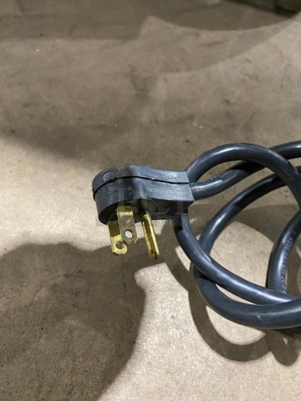 Close-up of a black power cord with a three-prong plug, featuring slight wear, resting on a concrete surface.