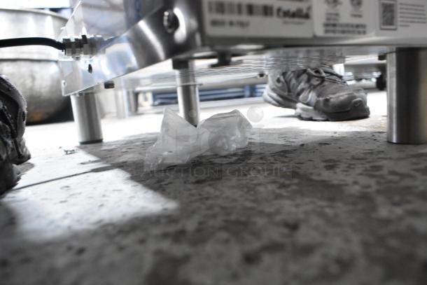 A stainless steel Estella 348PEM4 countertop pasta extruder machine sits on a floor, viewed from a low angle, showing its base and power connection.