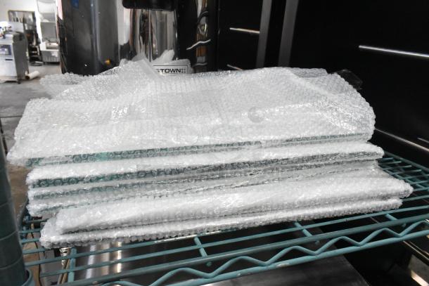 Stack of 6 new scratch and dent glass panes wrapped in bubble wrap on a metal rack, indicating delicate handling.