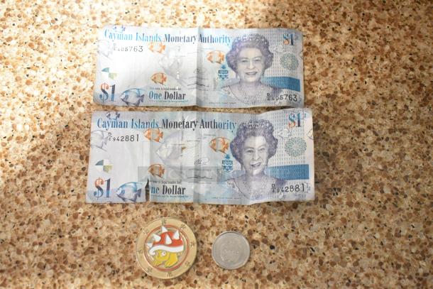Four items: two Cayman Islands $1 bills, a Super Mario coin, and an Apple coin on a speckled surface.