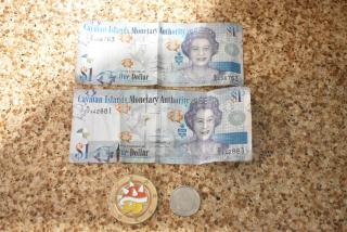 Four items: two Cayman Islands $1 bills, a Super Mario coin, and an Apple coin on a speckled surface.