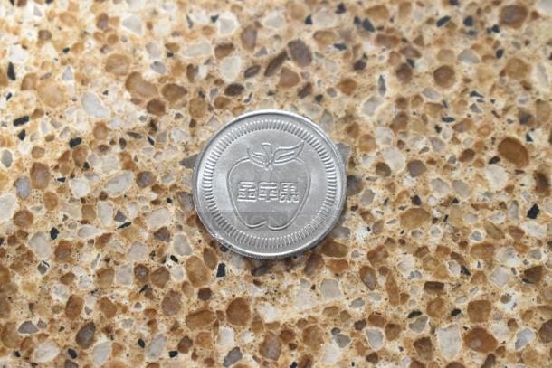 Apple coin with embossed apple and text, shown on textured surface; part of a four-item auction lot including two $1 Cayman Islands notes and a Super Mario coin.
