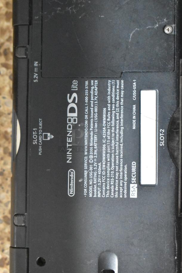 Nintendo DS Lite with visible tags; includes model and compliance details. Black casing, signs of wear.