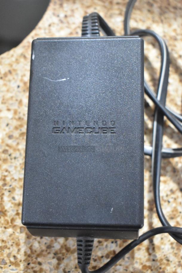 Nintendo GameCube power adapter, black, with cable. Shows minor wear, text "NINTENDO GAMECUBE" visible.