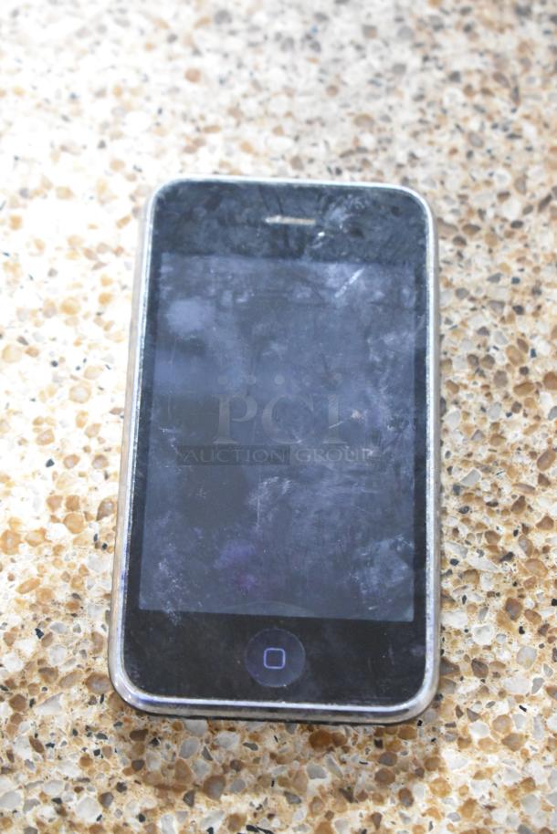 Apple iPhone A1303 with visible scratches on screen, placed on a pebble-patterned surface.