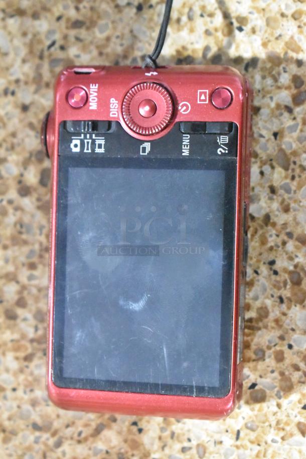 Red Sony Cyber-shot digital camera, back view, showing screen and controls, slightly scratched, on speckled surface.