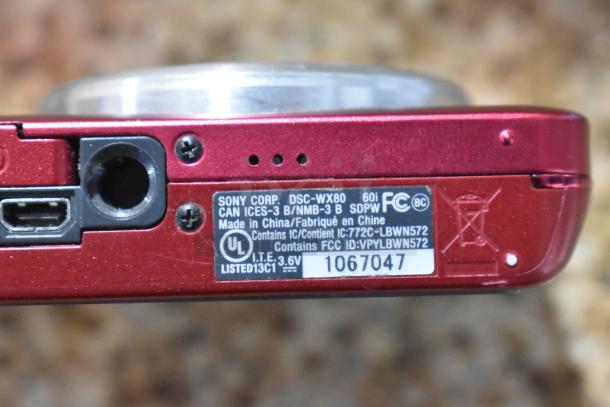 Red Sony Cyber-shot DSC-WX80 digital camera base with ports and model label, showing FCC and UL markings.