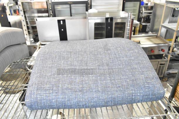 4 brand new scratch and dent seat cushions, blue-gray fabric, lightly textured, displayed on a metal shelf in a warehouse setting.
