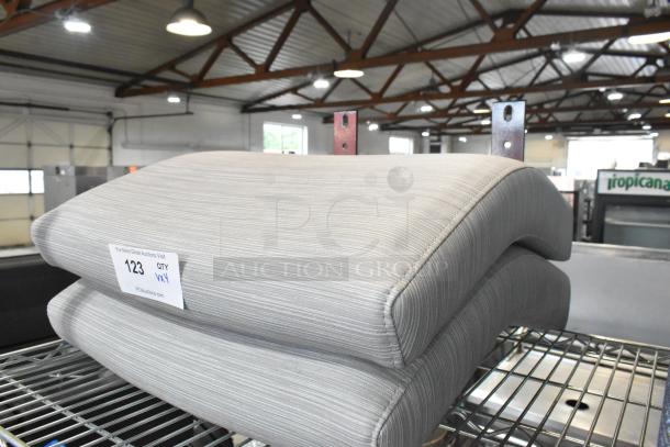 Four brand new scratch and dent seat cushions, grey striped fabric, stacked on metal shelf, auction tag reads "123 QTY VXY".