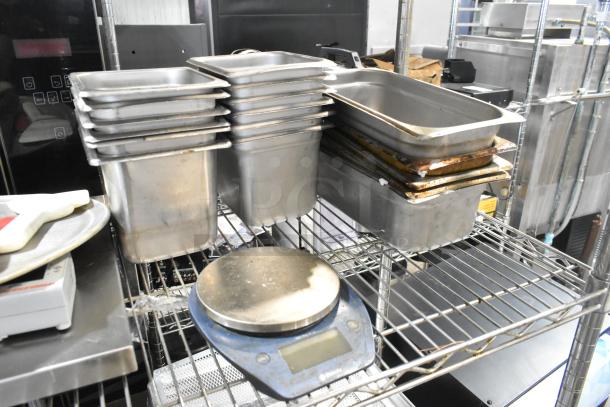 Lot of stainless steel drop-in bins and utensils on a wire shelf, including various-sized bins and a digital kitchen scale.