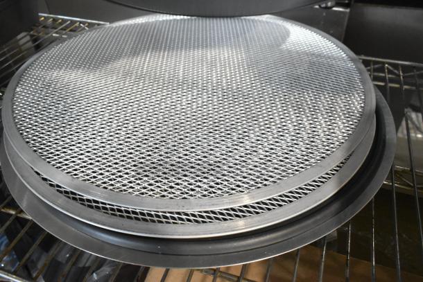 18 scratch and dent metal mesh pans and round perforated baking sheets, new condition, stacked on a metal rack.