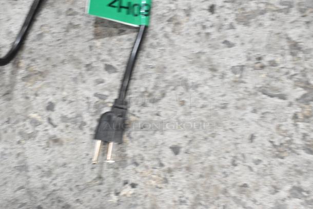 Blurred image showing a power cord with a standard plug, partially obscured green tag in background.