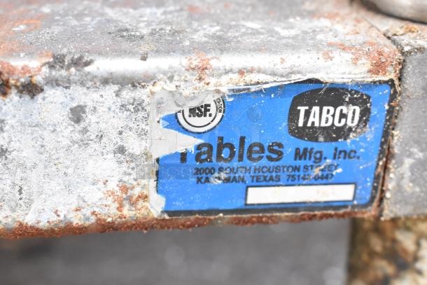 Stainless steel equipment stand, showing rust and wear, with a label from Tables Mfg. Inc., featuring commercial casters.