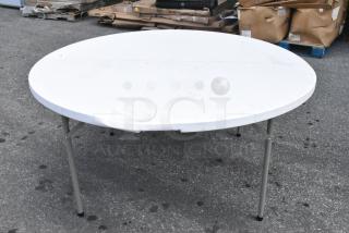 White round folding table with metal legs on outdoor pavement. Slight wear visible, ideal for flexible seating.