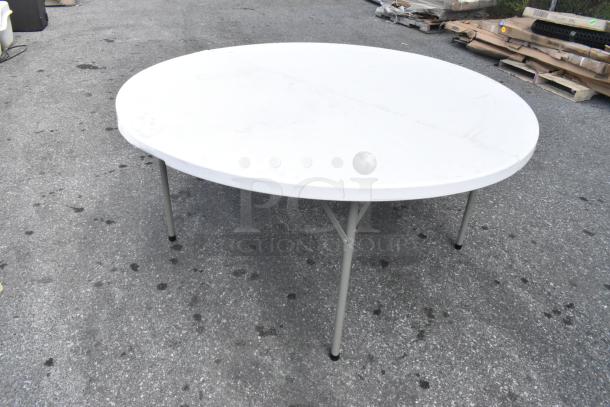 White round folding table on a gray surface, showing slight wear. Metal legs, portable design. Ideal for events or gatherings.