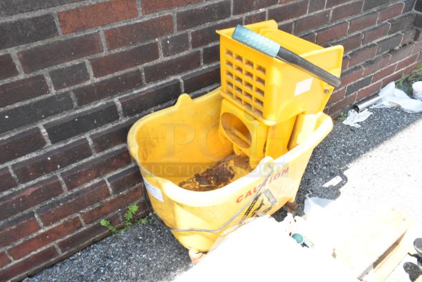 Yellow mop bucket with wringing attachment and commercial casters, labeled "CAUTION," shows signs of wear.