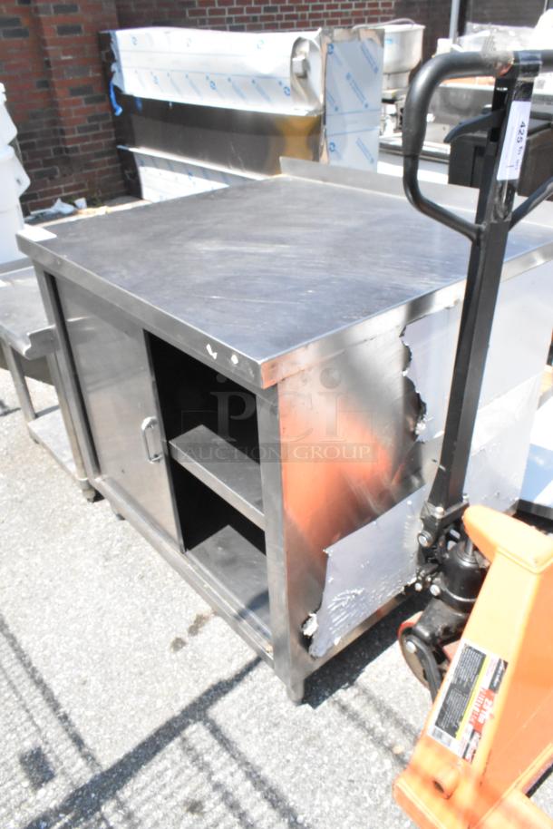 Stainless steel table with two under shelves and door, visible surface wear, and minor dents on side. Ideal for storage.