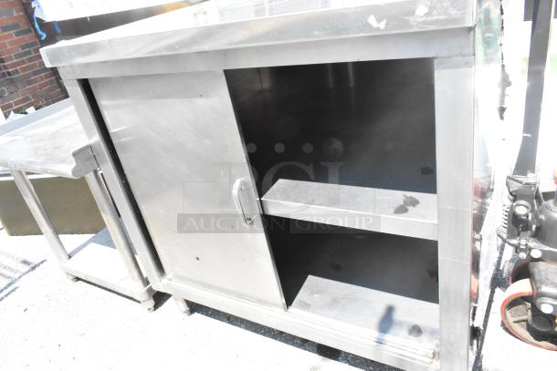 Stainless steel table with two under shelves and a door, showing signs of use, ideal for kitchen or workshop storage.