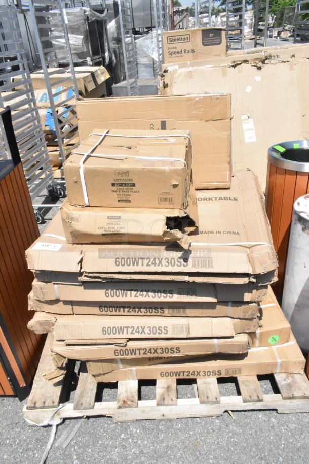 Stack of boxed commercial kitchen items, including 7 Regency 24"x30" stainless steel work tables and Lancaster cast iron table base plates, some boxes are scratched and dented.