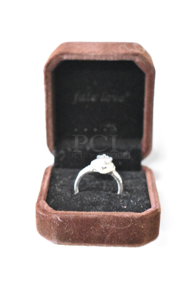 Alt text: Silver ring with a clear stone in a brown velvet jewelry box. Box lined with black fabric, subtle wear visible.