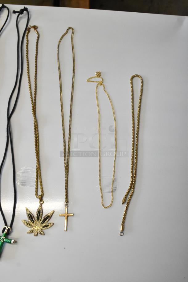 Lot of jewelry including necklaces with leaf and cross pendants, gold chains, black cords, laid out on white surface.
