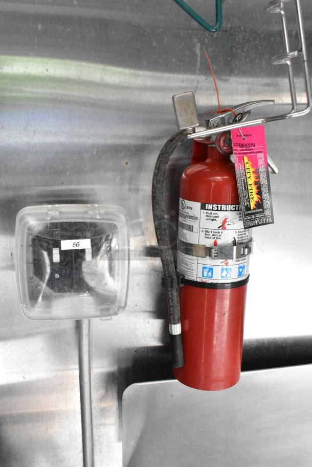 Red fire extinguisher with instructional label and inspection tag, mounted inside stainless steel food trailer, includes outlet cover.