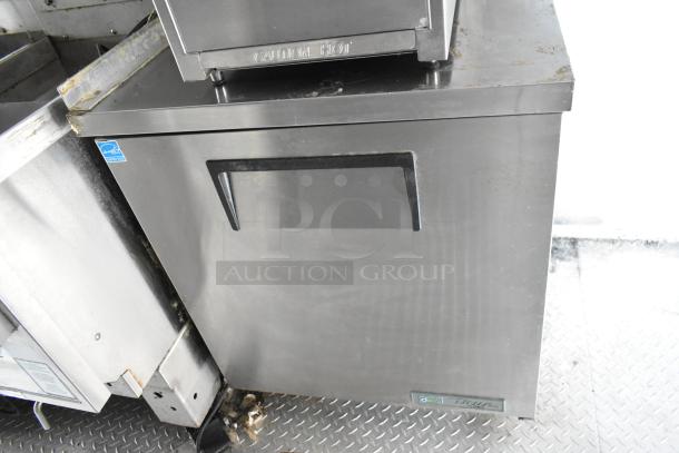 Food trailer stainless steel under-counter fridge. Features "True" branding and "CAUTION HOT" label. Exhibits minor wear.